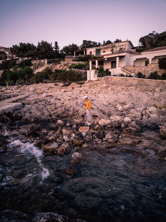 A person in an orange jacket explores rocky seaside cliffs at twilight near coastal houses.