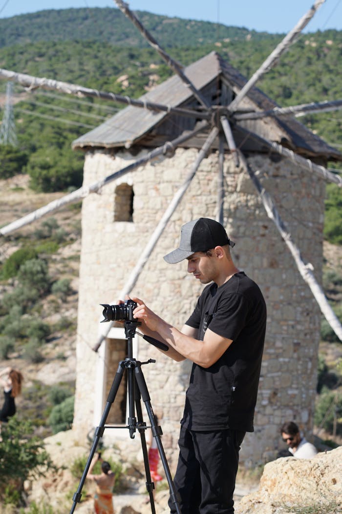 A man operates a camera on a tripod near an old stone windmill on a sunny day.
